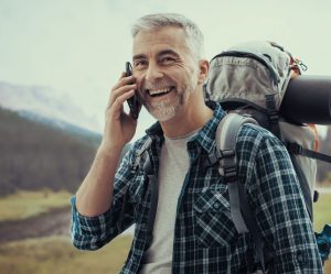 Bergwanderer mit grauen Haaren mit Rucksack spricht fröhlich in sein Handy.